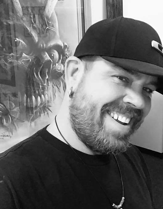 Ron, wearing a black cap and shirt with a beard and an earring, smiles warmly. In the background at Till Death Tattoo Studio near Lakeland, a large artwork featuring a skull with horns is prominently displayed.