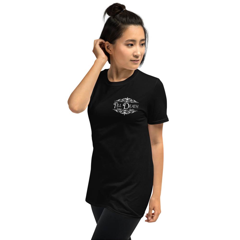A person in a unisex basic softstyle t-shirt, black and short-sleeved, featuring "Till Death" printed on the front, stands with one hand touching their hair.
