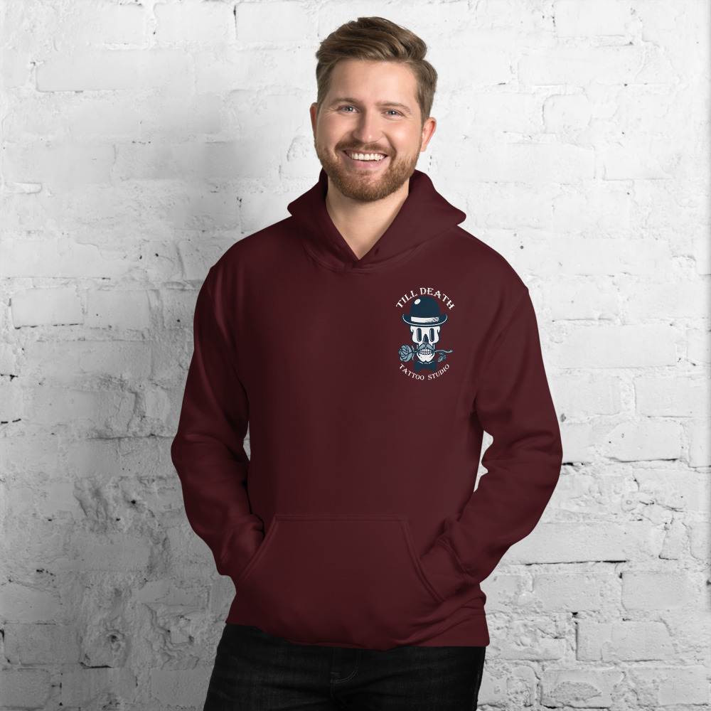 A bearded man smiles while wearing the Unisex Heavy Blend Hoodie in maroon, displaying a skull graphic and the text "Till Death Tattoo Studio," set against a white brick wall.