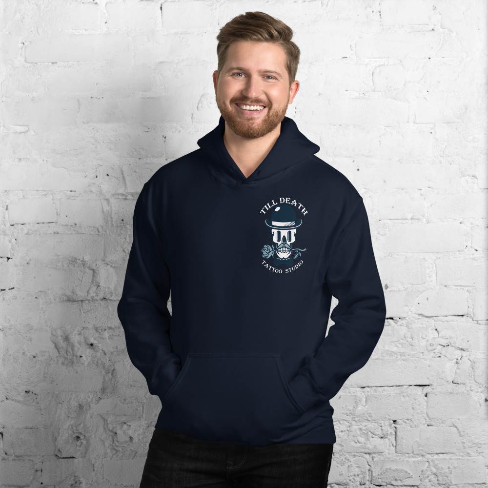 A man wearing a navy unisex heavy blend hoodie, featuring the "Till Death Tattoo Studio" logo, stands smiling against a white brick wall.