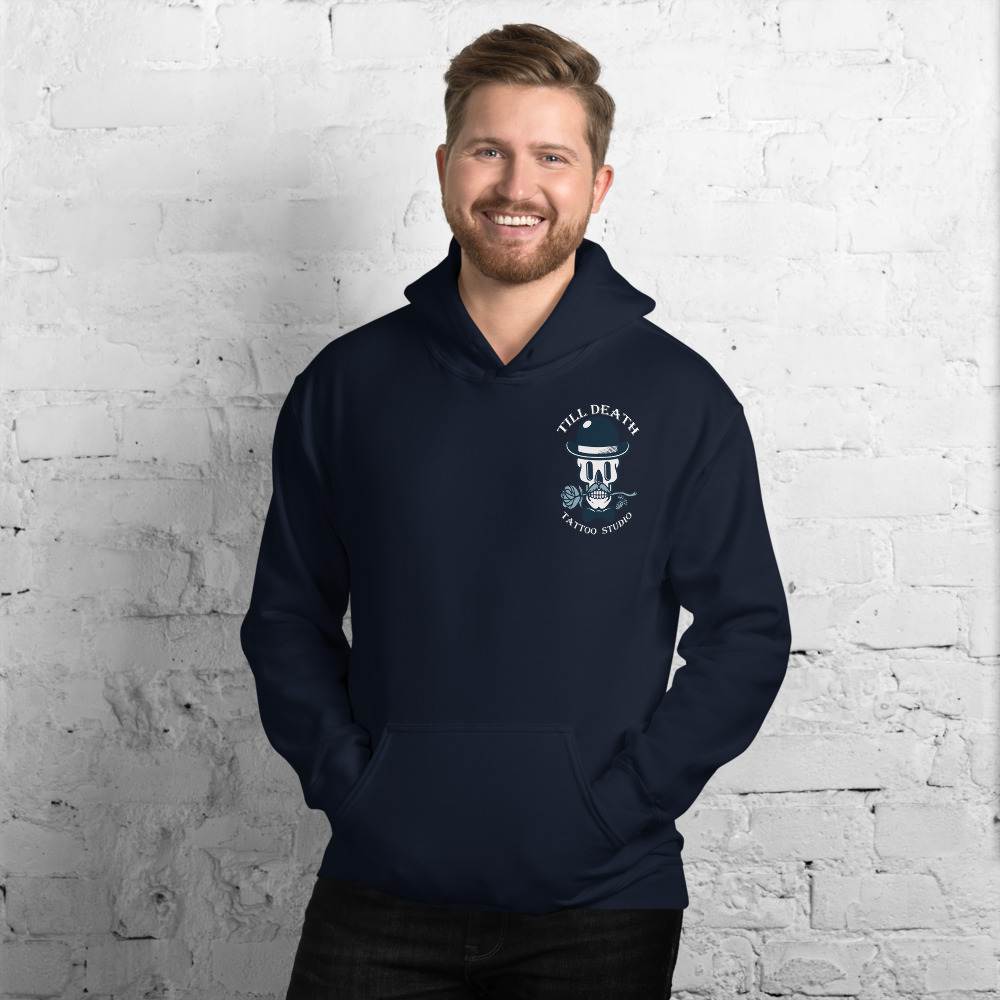 A man wearing a navy unisex heavy blend hoodie stands smiling against a white brick wall.