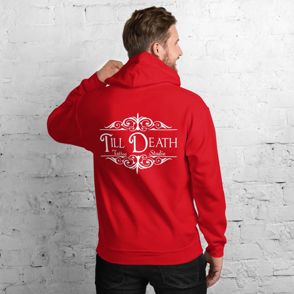 A man confidently stands against a white brick wall, wearing a unisex heavy blend hoodie in a vibrant red color, featuring "Till Death Tattoo Studio" prominently displayed on the back.