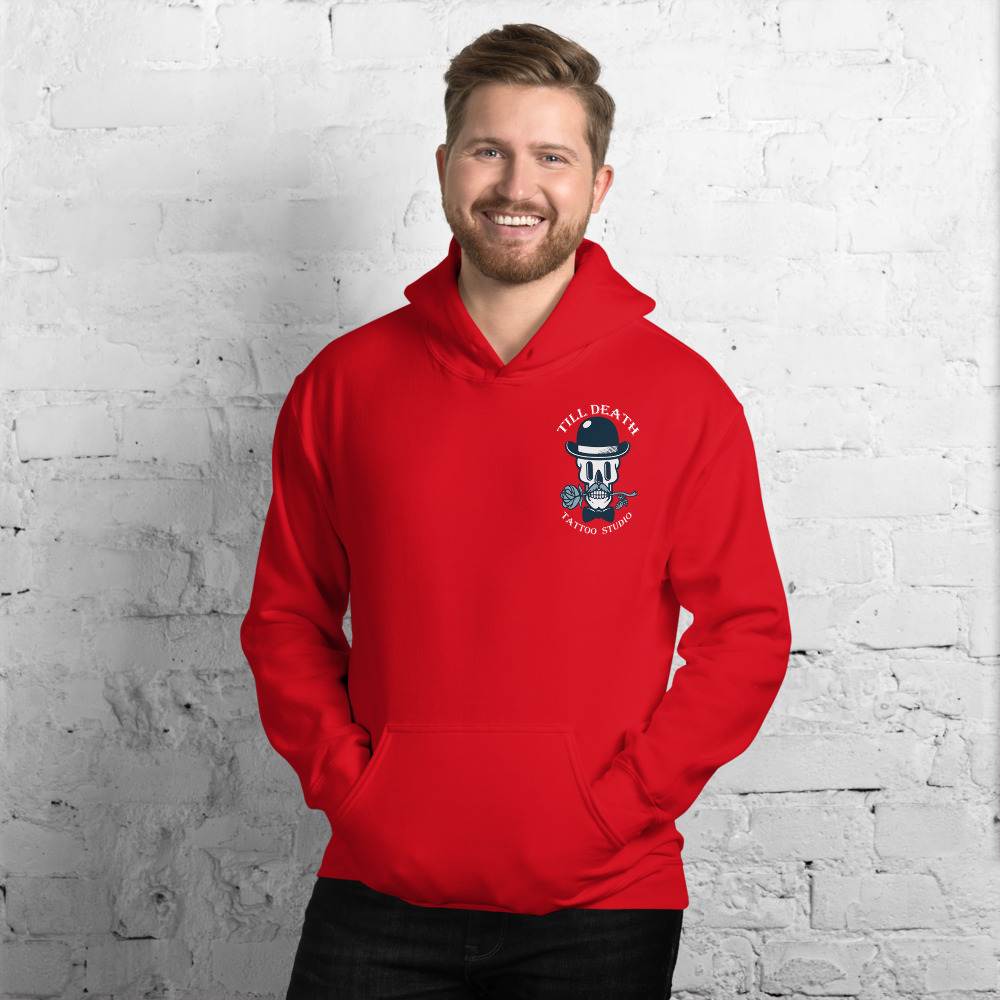 A person in a unisex heavy blend red hoodie, featuring a skull design, stands against a white brick wall.