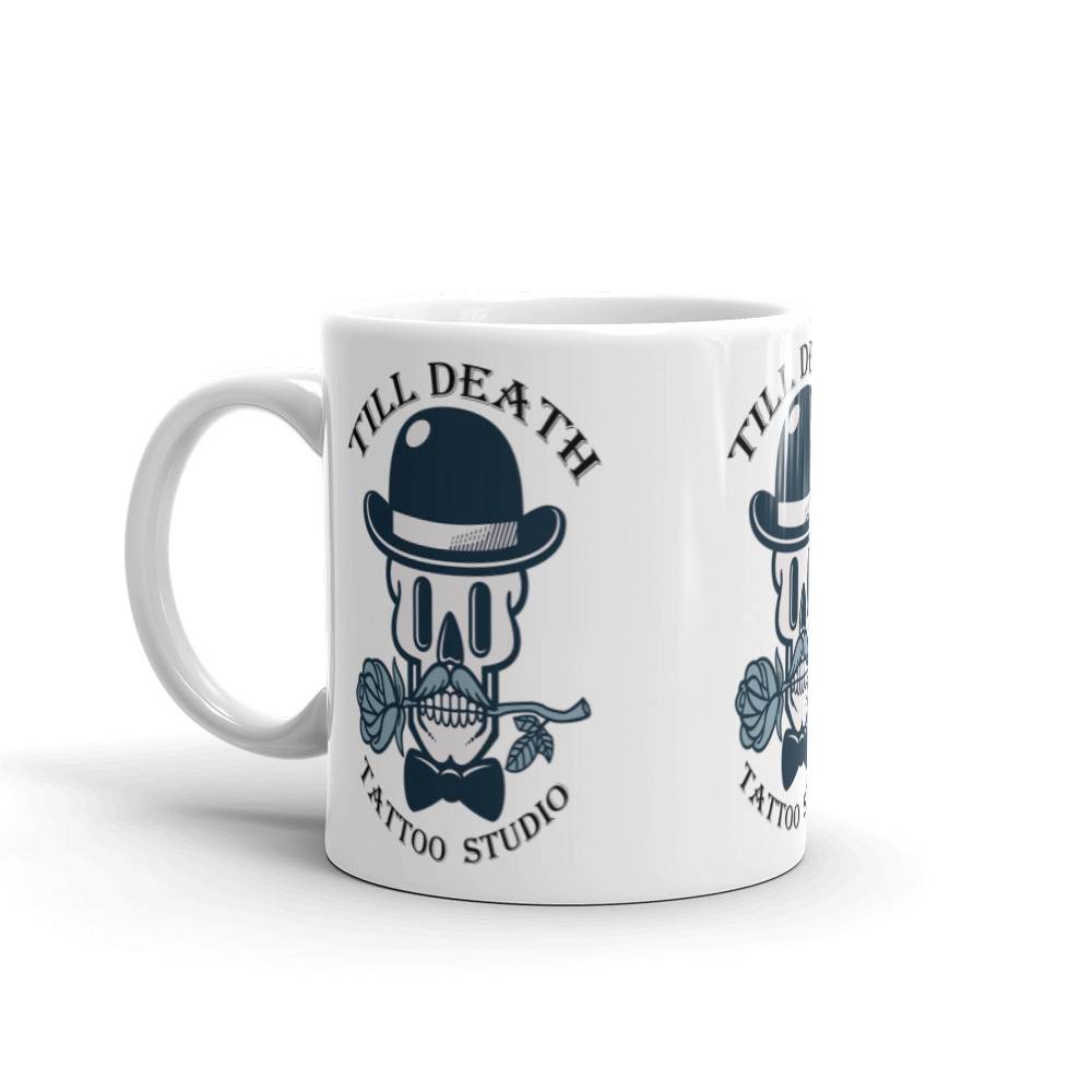 The white glossy mug (11 oz) showcases a cartoon skull wearing a bowler hat and holding a rose, with "Till Death Tattoo Studio" elegantly presented on it.