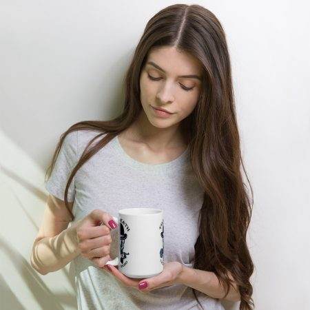 A woman with long hair holds a white glossy 15oz mug, looking down. She is wearing a grey t-shirt and stands as though waiting in a tattoo studio, against a plain white background.