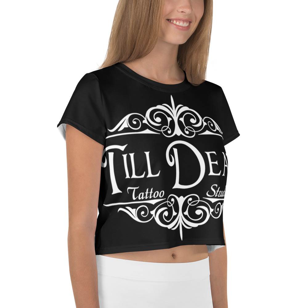 A woman in a chic all-over-print crop tee showcases "Till Death Tattoo Studio" in bold white text, embellished with detailed decorative designs, making it a fashionable addition to any wardrobe.