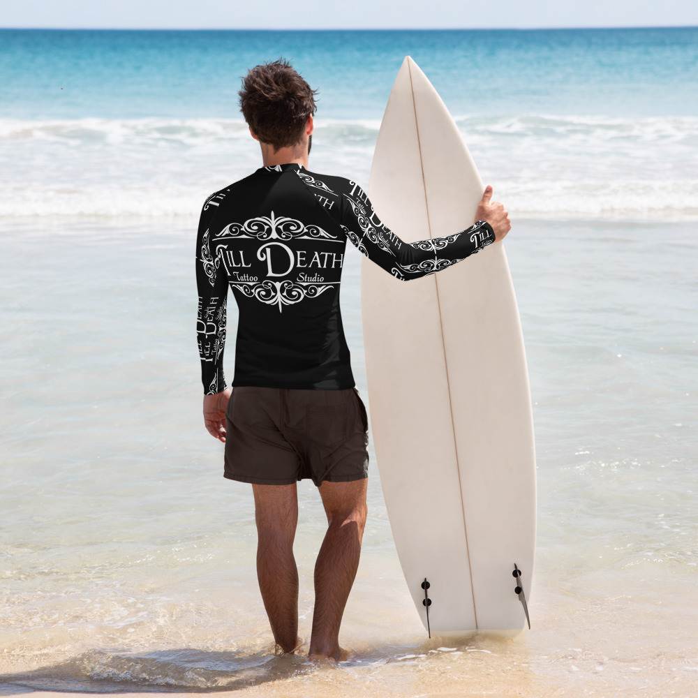 A person stands on the beach holding a surfboard, wearing an all-over-print men's rash guard with a white back and shorts, facing the ocean.