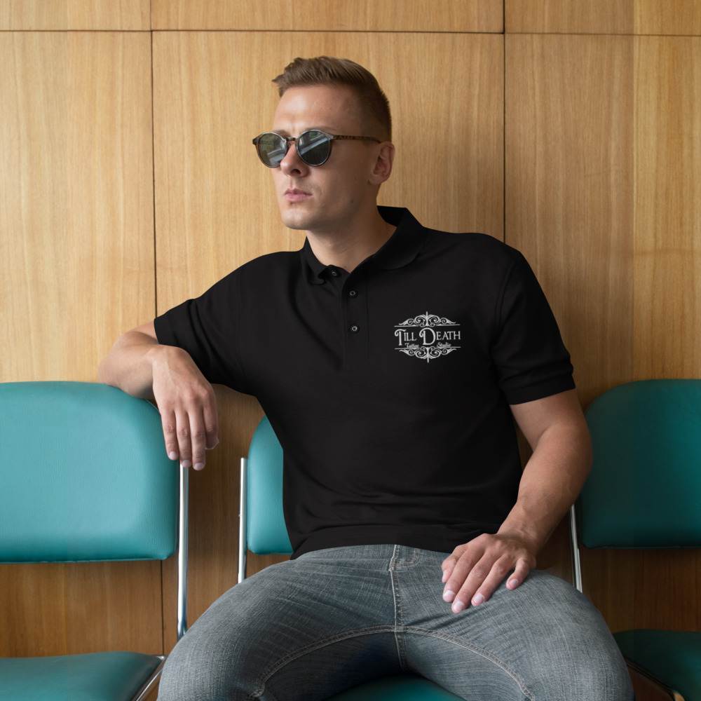 A person in sunglasses wearing the "premium-polo-shirt-black" sits on a teal chair against a wood-paneled wall, evoking the chic vibe typical of a tattoo studio.