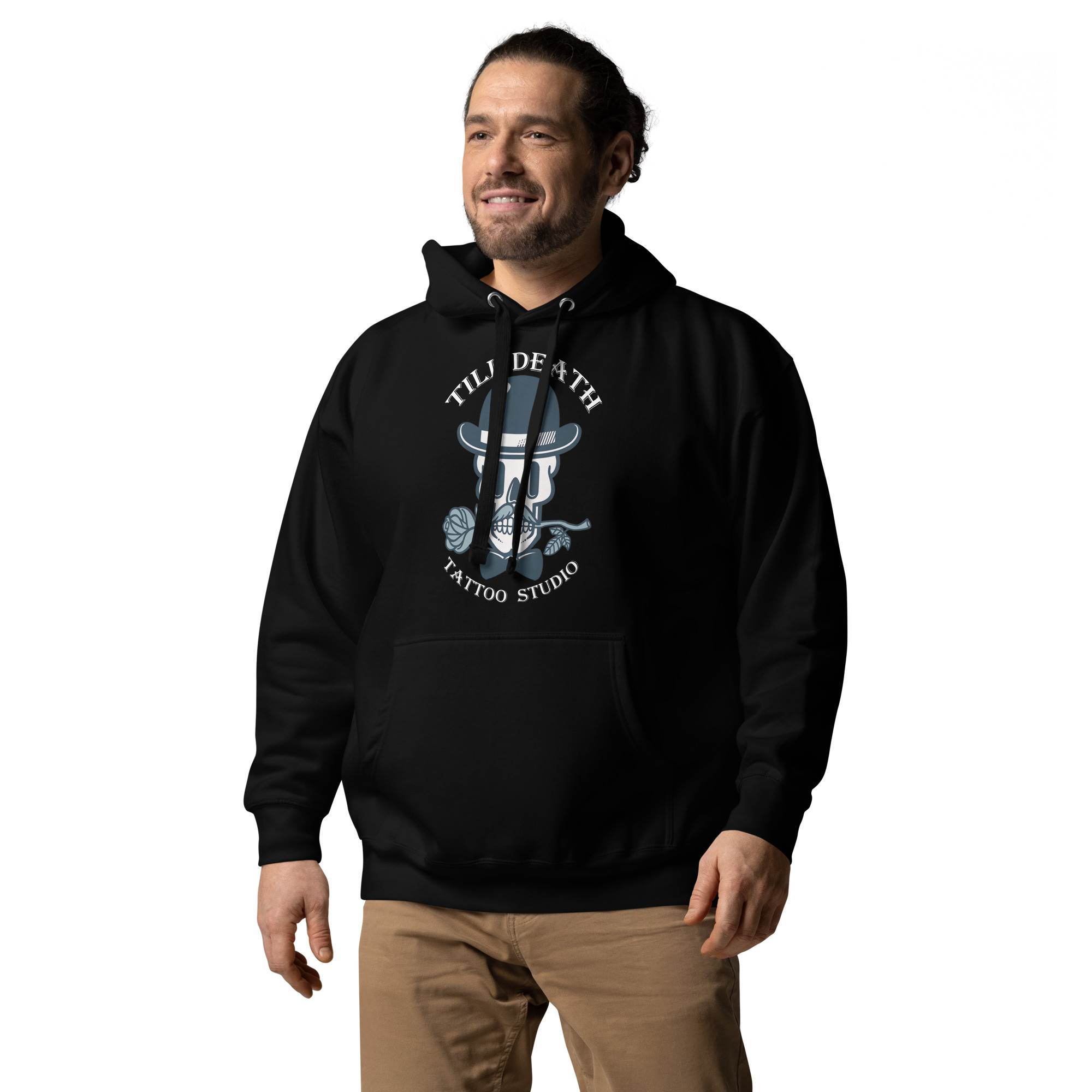 A person wearing a unisex premium hoodie in black, featuring the "Till Death Tattoo Studio" skull design, stands smiling, embodying the perfect fusion of art and style.