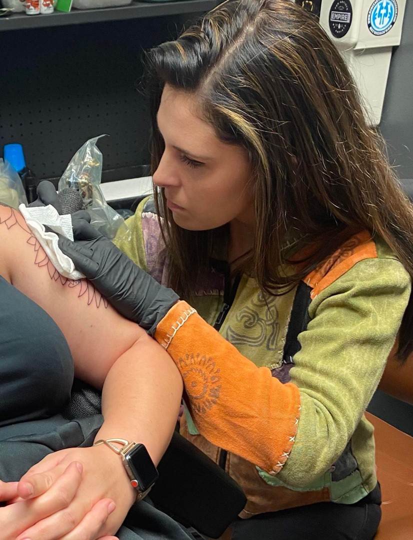 A woman with long brown hair is concentrating while tattooing a person's upper arm at Till Death Tattoo Studio, wearing a colorful jacket and black gloves. (Product Name: 361697532_605169218392728_9070277464491758014_n)