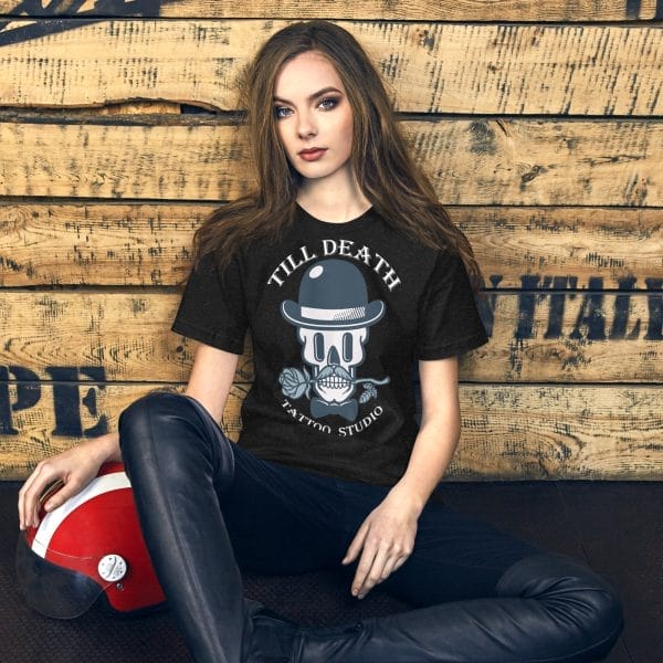 A woman sits against a wooden wall, wearing a black unisex staple t-shirt and leather pants, with a red helmet beside her.