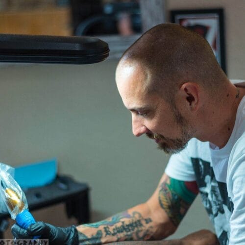 Bryan Skoltz, a tattoo artist with tattoos on his arms, meticulously works on a design at Till Death Tattoo Studio. Clad in a white shirt and gloves, he skillfully maneuvers the tattoo machine, embodying precision and artistry in every stroke.