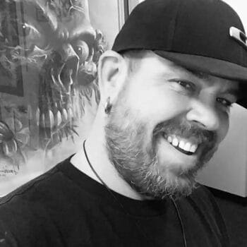 Ron, wearing a black cap and shirt with a beard and an earring, smiles warmly. In the background at Till Death Tattoo Studio near Lakeland, a large artwork featuring a skull with horns is prominently displayed.