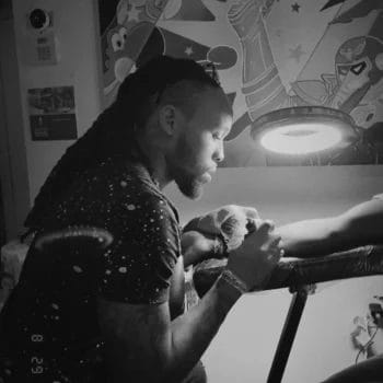 In a dimly lit room decorated with cartoon art, a man with long hair tattoos an arm beneath a lamp, captured in an image at Till Death Tattoo Studio, a renowned tattoo shop near Lakeland.